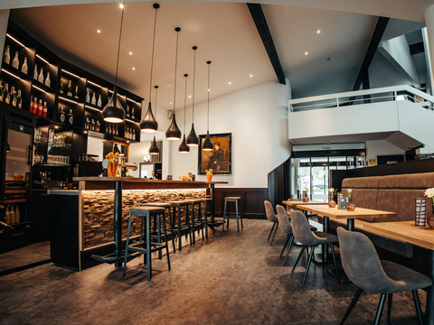 Mittendrin (3).jpg Modern eingerichtetes Café mit Steinbar, hölzernen Tischen, grauen Stühlen und stimmungsvoller Beleuchtung.