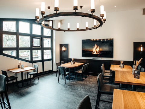 Mittendrin (6).jpg Modern eingerichtetes Restaurant mit großen Fenstern, Holztischen, stilvollen Leuchten und Kaminbild.