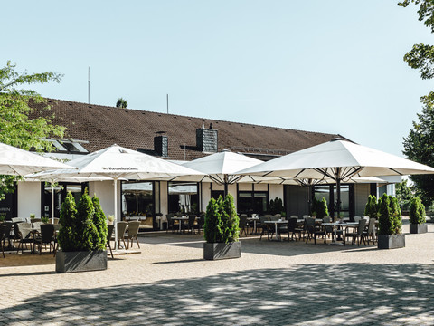 Terrasse 1.jpg Große Terrasse eines Restaurants mit weißen Sonnenschirmen, Tischen und Stühlen, umgeben von Pflanzen.