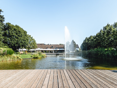 Weiher 2.jpg Holzdeck am Teich mit Springbrunnen, umgeben von Bäumen und einem Gebäude im Hintergrund.