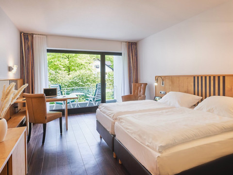 PHN_Comfort_Doppelzimmer_Vinyl_QF.jpg Helles Hotelzimmer mit zwei Einzelbetten, großen Fenstern und Balkon mit Blick auf grüne Landschaft.
