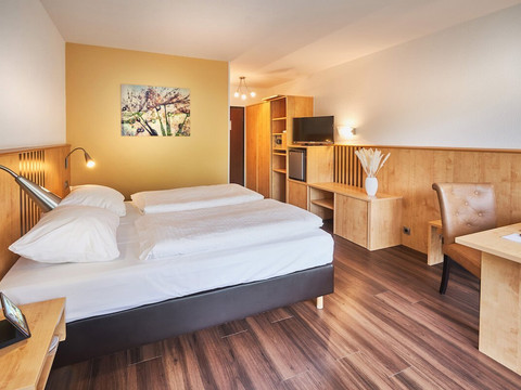 PHN_Comfort_Doppelzimmer_Vinyl_QF2.jpg Modernes Hotelzimmer mit Doppelbett, Holzmöbeln und Wandbild, einladend dekoriert, gut beleuchtet.