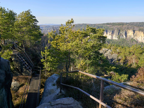Blick von einem Waldweg mit Geländer auf eine felsige Schlucht und bewaldete Landschaft.