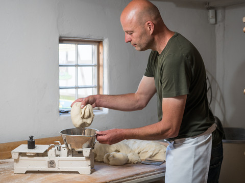 Backhaus aus Overath-Kepplerburg Ein Bäcker im grauen Shirt formt Teig in einer traditionellen Backstube mit einer alten Waage.