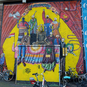 Mural Das Bild zeigt ein farbenfrohes Wandgemälde in Köln mit stilisierten Figuren, die Kleidung in leuchtenden Farben tragen. Fahrräder sind vor der gemalten Wand geparkt.The picture shows a colourful mural in Cologne with stylized figures wearing clothes in bright colors. Bicycles are parked in front of the painted wall.