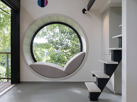 Innenansicht Baumhaus Rundes Fenster mit integrierter Sitzgelegenheit bietet Ausblick auf grüne Bäume und modernen Innenraum.
