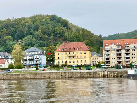 Fahrradbox neben dem Rathaus Uferansicht mit einem gelben und einem blauen Gebäude vor einem bewaldeten Hügel, ruhige Atmosphäre, grauer Himmel.Shore view with a yellow and a blue building in front of a wooded hill, calm atmosphere, gray sky.Pohled na pobřeží se žlutou a modrou budovou před zalesněným kopcem, klidná atmosféra, šedá obloha.Widok na brzeg z żółtym i niebieskim budynkiem przed zalesionym wzgórzem, spokojna atmosfera, szare niebo.Kustgezicht met een geel en een blauw gebouw voor een beboste heuvel, rustige sfeer, grijze lucht.Vista della costa con un edificio giallo e uno blu di fronte a una collina boscosa, atmosfera tranquilla, cielo grigio.