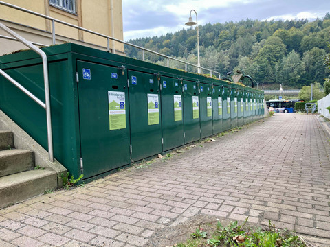 Fietsdozen_Bad_Schandau Grüne Fahrradgaragen mit Informationsplakaten stehen entlang eines gepflasterten Weges, umgeben von Bäumen und einem Bus im Hintergrund.