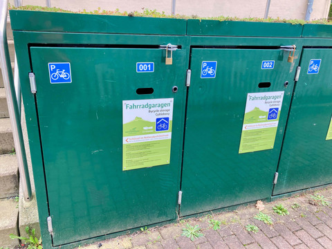 Fietsgarage_Bad_Schandau Grüne Fahrradgaragen mit blauen Fahrrad-Piktogrammen und Nummern 001 und 002, auf einem gepflasterten Boden vor einer Treppe.
