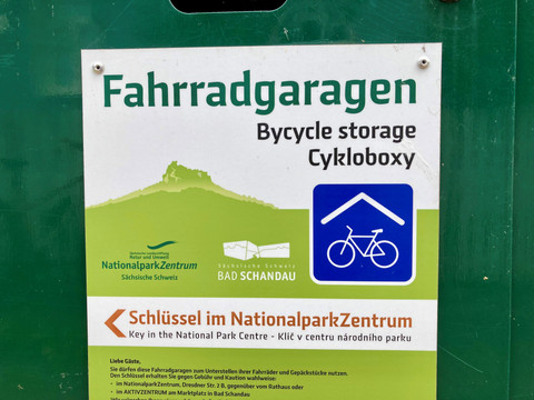 Fietsgarage_Bad_Schandau Schild mit der Aufschrift "Fahrradgaragen" in drei Sprachen, darunter ein Symbol für Fahrradaufbewahrung und Hinweise zur Schlüsselausgabe im Nationalparkzentrum.