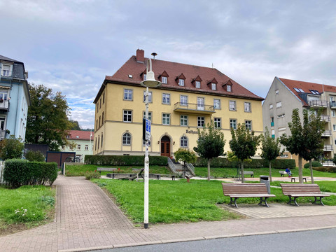 Toegang_tot_de_fiets_garages_van_uit_de_Lbe Ein gelbes Rathaus mit rotem Dach steht zwischen Bäumen und Bänken in einem grünen Park; der Himmel ist bewölkt.