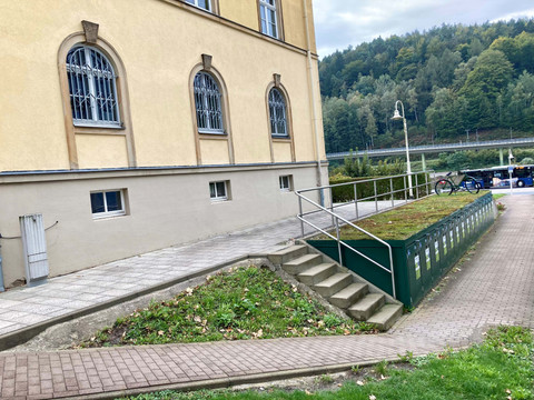 Fietsdozen_Bad_Schandau Gelbes Gebäude mit vergitterten Fenstern, daneben ein Fahrrad auf einer Rampe. Im Hintergrund sind Bäume und ein Bus zu sehen.