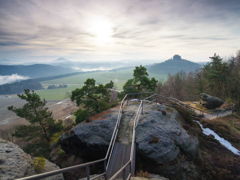Ausblick von der Kaiserkrone Steinpfad mit Geländer führt über Felsen, umgeben von Bäumen und Wiesen, im Hintergrund neblige Hügel unter bewölktem Himmel.