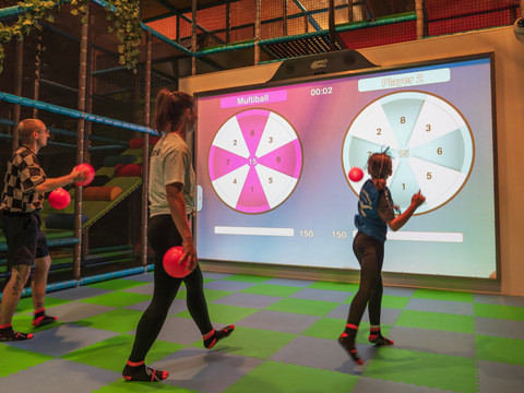 Multiball Drei Personen spielen Multiball an einem interaktiven Bildschirm auf einem bunten Hallenboden.Three people play multiball on an interactive screen on a colorful hall floor.Tři osoby hrají multiball na interaktivní obrazovce na podlaze barevné haly.Trzy osoby grają w multiball na interaktywnym ekranie na kolorowej podłodze hali.Drie mensen spelen multiball op een interactief scherm op een kleurrijke zaalvloer.Tre persone giocano a multiball su uno schermo interattivo sul pavimento di una sala colorata.