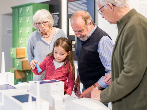 Am Fabrikmodell Eine Gruppe älterer Menschen und ein Kind betrachten gemeinsam ein architektonisches Modell in einem Museum.
