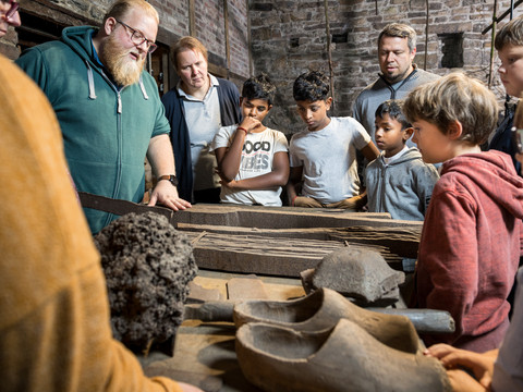 Oelchenshammer Gruppe von Kindern und Erwachsenen betrachtet Werkzeuge auf einem Holztisch in historischer Werkstatt.