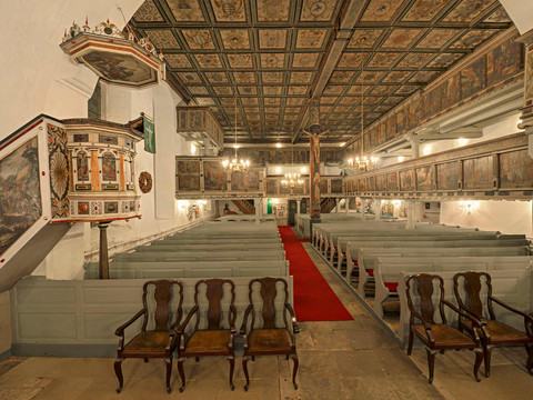 Vstupní prostor evangelického luteránského kostela Historische Kirche mit bemalter Holzdecke, Kanzel und Bänken; rote Teppichläufer und hölzerne Stühle schaffen eine feierliche Atmosphäre.