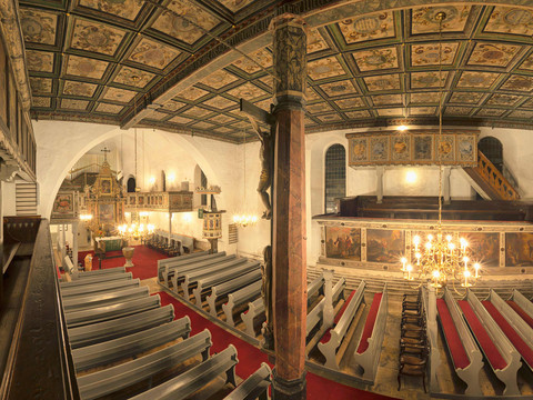 Pohled na interiér z galerie Innenansicht einer Kirche mit bemalter Holzdecke, roten Teppichen und grauen Holzbänken; ein Kronleuchter hängt zentral.