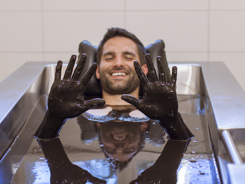 Naturmoorvollbad Mann genießt entspannendes Bad in dunklem Naturmoor, während er lächelnd seine Hände zeigt.Man enjoying a relaxing bath in dark natural mud while smiling and showing his hands.Muž si užívá relaxační koupel v tmavém přírodním bahně, usmívá se a ukazuje si ruce.Mężczyzna cieszy się relaksującą kąpielą w ciemnym naturalnym błocie, uśmiechając się i pokazując ręce.Man geniet van een ontspannend bad in donkere natuurlijke modder terwijl hij glimlacht en zijn handen laat zien.Uomo che si gode un bagno rilassante nel fango naturale scuro mentre sorride e mostra le mani.