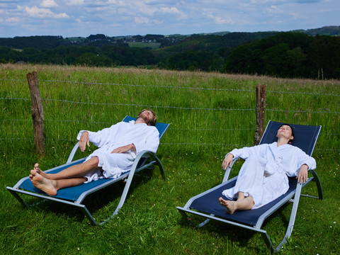 Trans World Hotel Kranichhöhe Zwei Personen entspannen in weißen Bademänteln auf Liegestühlen vor einer grünen Wiese und bewölktem Himmel.