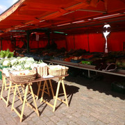 Stand auf dem Aper Wochenmarkt Ein Marktstand mit frischen Gemüsekisten unter orangefarbenen Planen auf einem gepflasterten Platz.