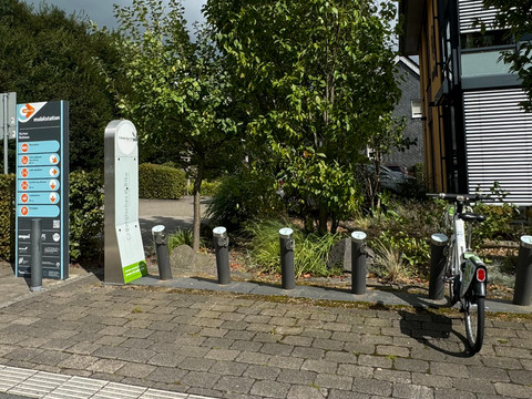 E-Bike Verleihstation Fahrradparkplätze mit Ladesäulen, ein Fahrrad steht bereit, umgeben von Bäumen und einem modernen Gebäude.