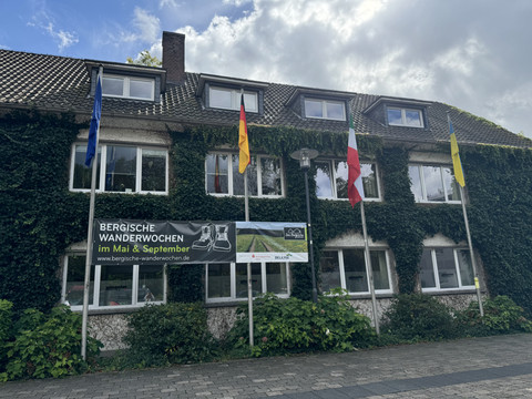 Karlheinz-Stockhausen Platz - Banner Wanderwochen Ein mit Efeu bewachsenes Gebäude mit Bannern und Fahnen, die auf das Bergische Wanderwochen-Event hinweisen.