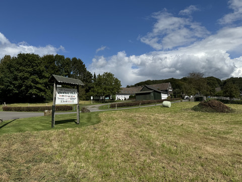 Landhaus Fuchs - Ansicht von Hauptstraße aus Ländliche Szene in Kürten mit grünem Feld, bewaldeten Hügeln und einem Schild vor Bauernhof.