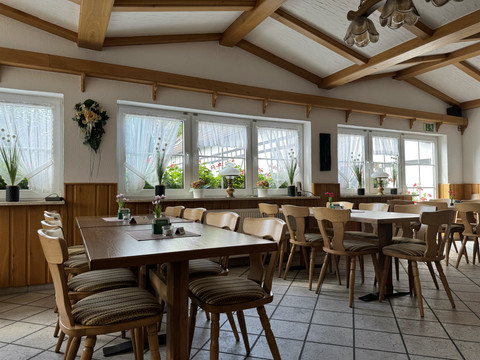 Landhaus Fuchs - Ansicht Innenbereich Ein gemütlicher Gastraum mit Holzmöbeln und Tischdekoration, umgeben von großen, blumengeschmückten Fenstern.