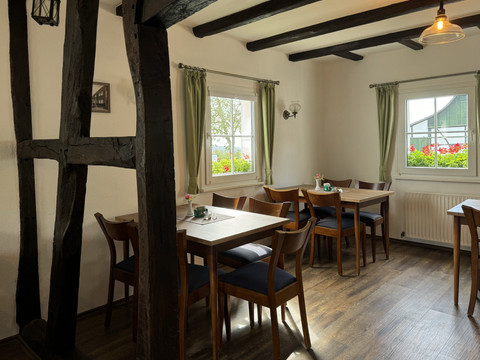 Landhaus Fuchs - Ansicht Innenbereich Gemütliches Café mit Holzbalken, hellen Wänden und großen Fenstern mit Sicht auf blühende Gärten.