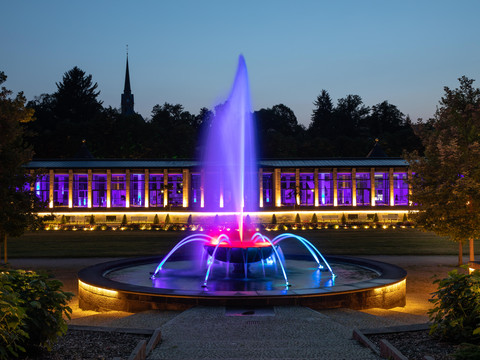 Springbrunnen Erlebnis und Lichterwelt Springbrunnen mit bunter Beleuchtung vor beleuchtetem Gebäude bei Abenddämmerung.Fountain with colorful lighting in front of an illuminated building at dusk.Fontána s barevným osvětlením před osvětlenou budovou za soumraku.Fontanna z kolorowym oświetleniem przed oświetlonym budynkiem o zmierzchu.Fontein met kleurrijke verlichting voor een verlicht gebouw in de schemering.Fontana con luci colorate davanti a un edificio illuminato al tramonto.