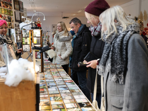 Weihnachtsmarkt Schloss Eulenbroich Menschen stöbern in einem Geschäft durch Postkartenauslagen, umgeben von winterlichen Bekleidungen.