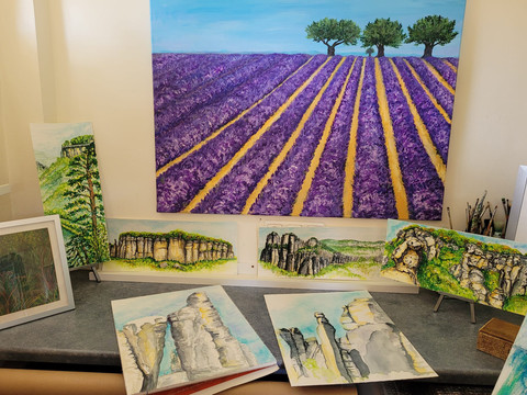 Auswahl Motive Andrea Moliere Mehrere Gemälde auf einem Tisch, darunter ein großes Bild eines violetten Lavendelfeldes mit Bäumen und kleinere Landschaftsmotive mit Felsen und Wäldern.