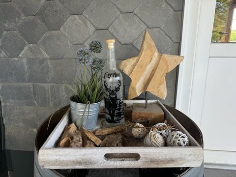 Jägerhof - Ansicht Deko Dekoratives Tablett auf einem Fass mit Flasche, Holzstern, Pflanzen und Deko-Obstkugeln.