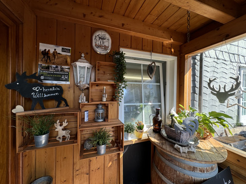 Jägerhof - Ansicht Tür Innenhof Ein einladender, rustikaler Holzveranda-Bereich mit Pflanzen, Laterne, Dekorationen und herzlichem Willkommensschild.