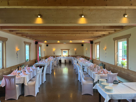 Jägerhof_Veranstaltungsraum.jpg Großer Festsaal mit weiß gedeckten Tischen, dekorierten Stühlen und Holzbalkendecke, bereit für Feier.