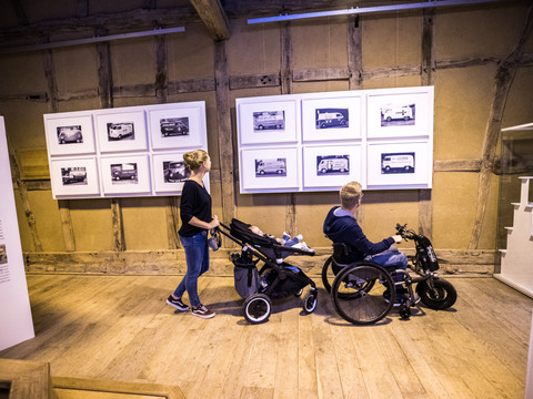 LVR-Freilichtmuseum Lindlar Menschen schauen in einer Ausstellung auf Fotos an der Wand. Einer der Besucher sitzt im Rollstuhl.