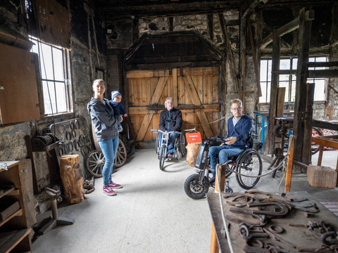 LVR-Freilichtmuseum Lindlar Menschen in einer historischen Schmiede, zwei im Rollstuhl, eine Person mit Kind, barrierefreie Umgebung.