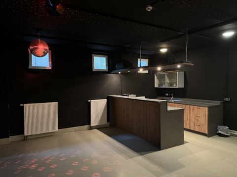 Jugendherberge Radevormwald - Partyraum Bar im dunklen Raum mit rotem Lichtschein, moderne Holztheke, Deckenstrahler und Fenster im Hintergrund.