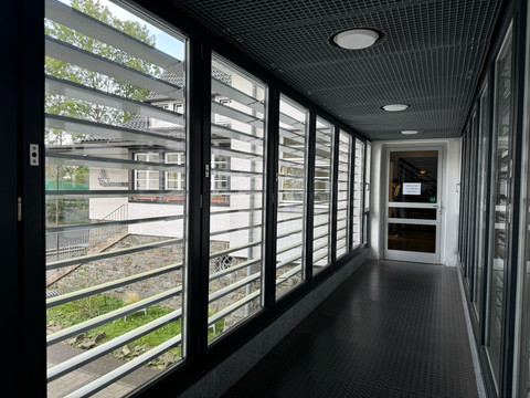 Jugendherberge Radevormwald - Ansicht Flur  Gang mit großen Fenstern und Lamellen, führt zu einer Glastür mit Hinweisschild. Links Grünfläche sichtbar.