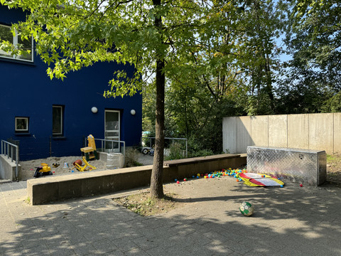 Jugendherberge Radevormwald - Ansicht Spielfläche Ein Spielplatz mit bunten Spielzeugen auf Betonfläche und umgeben von Bäumen und einem blauen Gebäude.