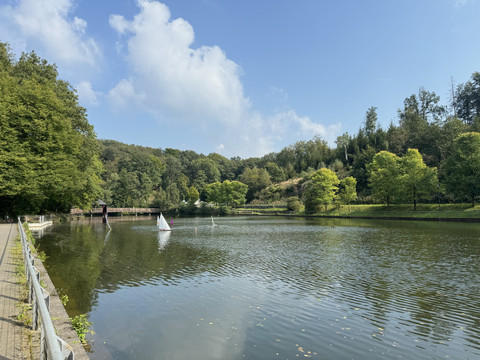 Café-Restaurant Uelfetal - Ansicht Uelfebad Ein malerischer See inmitten grüner Bäume mit Segelschiffen und einem steinernen Weg am Ufer.