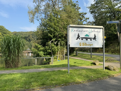 Café-Restaurant Uelfetal - Ansicht Schild  Schild am Wegesrand mit der Aufschrift "Restaurant Uelfetal", umgeben von grüner Natur und Bäumen.