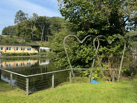 Café-Restaurant Uelfetal - Ansicht Uelfebad Metallskulptur in Herzform vor einem idyllischen See, umrahmt von Bäumen und einem gepflegten Rasen.