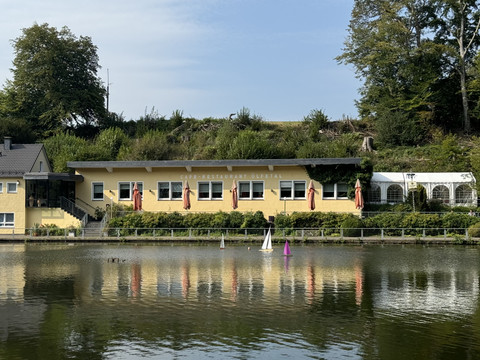Café-Restaurant Uelfetal - Außenansicht Gelbes Restaurant mit Terrasse und Sonnenschirmen am See, umgeben von Bäumen und Segelbooten.