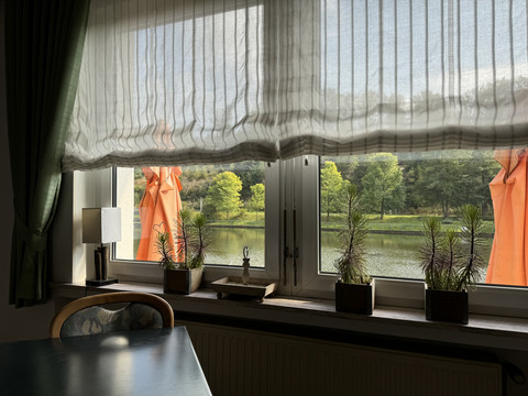 Café-Restaurant Uelfetal - Ansicht Fensterbank Blick durch ein Fenster mit weißen Vorhängen auf einen See, zwei orange Sonnenschirme und Bäume.