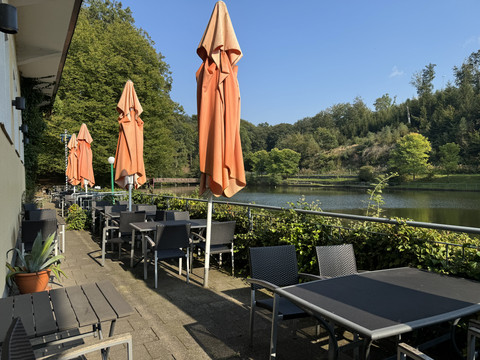 Café-Restaurant Uelfetal - Ansicht Außenterasse Terrasse eines Cafés mit roten Sonnenschirmen, Tischreihen, Blick auf einen ruhigen See und Bäume.