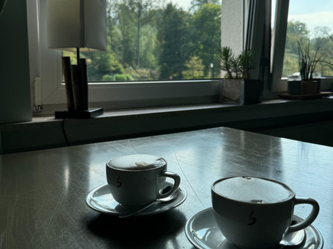 Café-Restaurant Uelfetal - Ansicht Cappuccino  Zwei Tassen Cappuccino auf einem Holztisch vor einem Fenster mit Blick auf grüne Bäume.