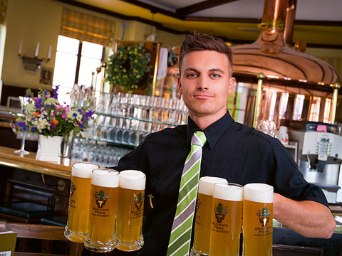 Brauhaus Pirna "Zum Gießer" Ein Kellner in schwarzem Hemd und gestreifter Krawatte trägt sechs Bierkrüge in einem gemütlichen Brauhaus mit Kupferkessel im Hintergrund.