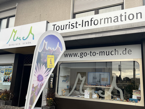 Tourist-Information  Tourist-Information Much mit Fensterschild und Fahne, darunter Blumenbeet, für Besucheranfragen.